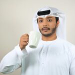 Young Middle Eastern man wearing traditional attire enjoys a cup of coffee indoors.